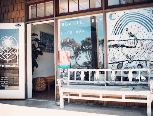 The Organic Tree storefront in Dana Point, CA. Picture shows an open door, a white bench to the right of the door, an artwork of the store window displays a negative print artwork of a person under a rainbow